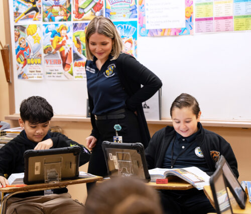 A teacher stands behind two students working from their textbooks