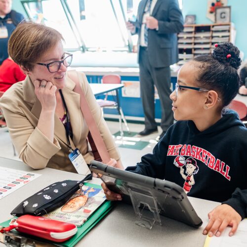 An educator sits with a young student a table on their tablet