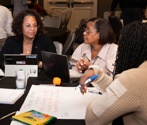 Three event attendees gather around a table discussion with laptops, markers, while taking notes on a large piece of paper