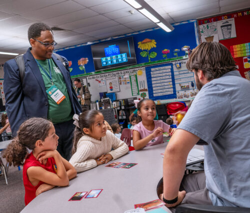 Three young students sit at a table in a classroom as an adult explains instructions