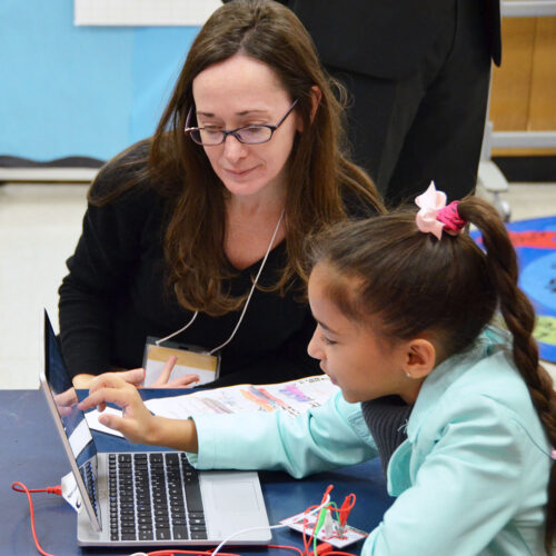 A teacher crouches next to a young student on their laptop