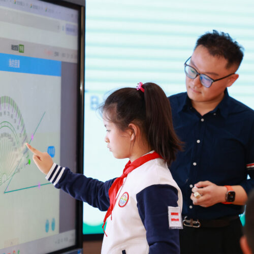 A young student touches a large interactive screen as a teacher stands next to her
