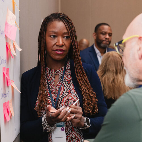 A systems leader holds a discussion with a colleague in front of sticky notes on the wall