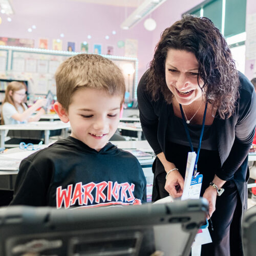 An adult leans down with an elementary student and smiles at a tablet in a classroom