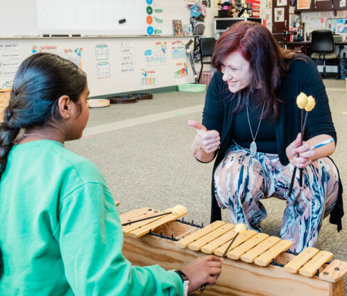 An instructor crouches down to the floor and gives a thumbs up to a young student playing the xylophone