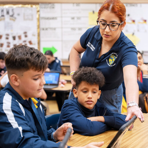 An instructor reaches over to help two students navigate their tablet in a classroom