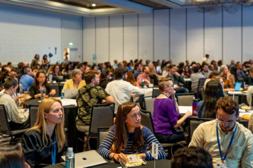 A crowded hotel ballroom of conference attendees seated at round tables, most turned attentively toward a speaker out of frame.