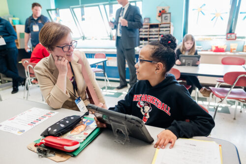 A female student in a black hoodie interacting with an female adult in business attire, they are engaged while the student holds a tablet