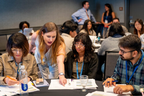 A facilitator leans over a conference table to point at a worksheet while three seated participants work through printed handouts.
