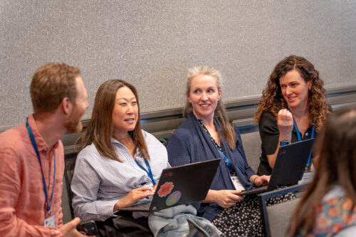A group of four people with laptops sit in chairs and talk and smile.