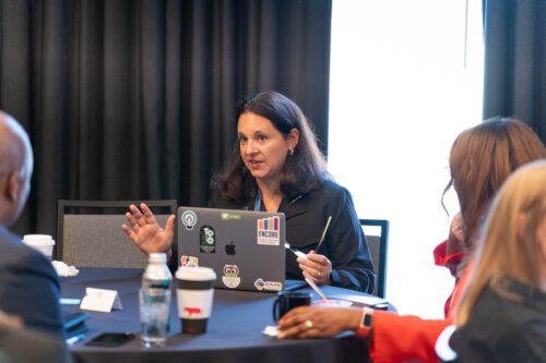 A woman speaks and gestures from behind a sticker-covered laptop as others listen at a round table in a conference session.