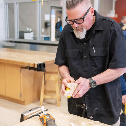 An older man wearing protective glasses sands a piece of wood in a workshop