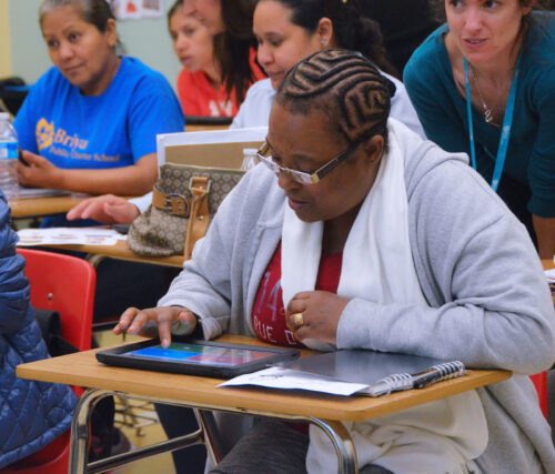 An adult sits at a desk in a classroom and interacts with their tablet