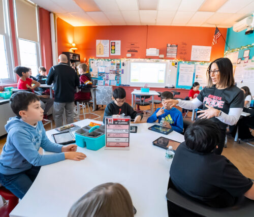 A teacher talks with a young elementary school student at a table in a colorful classroom