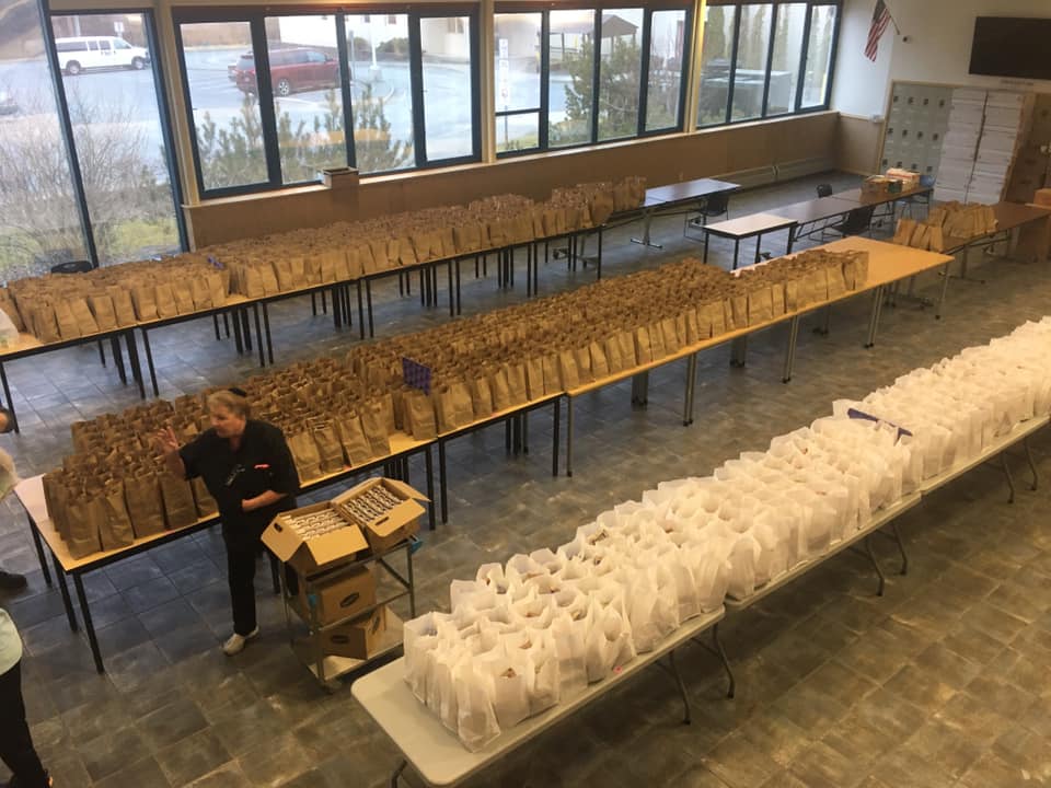 Three tables in a well-lit room with windows are lined with bags of meals to distribute the Sitka community