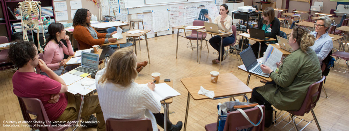 Photo of teachers sitting in desks in a circle looking at one another and speaking