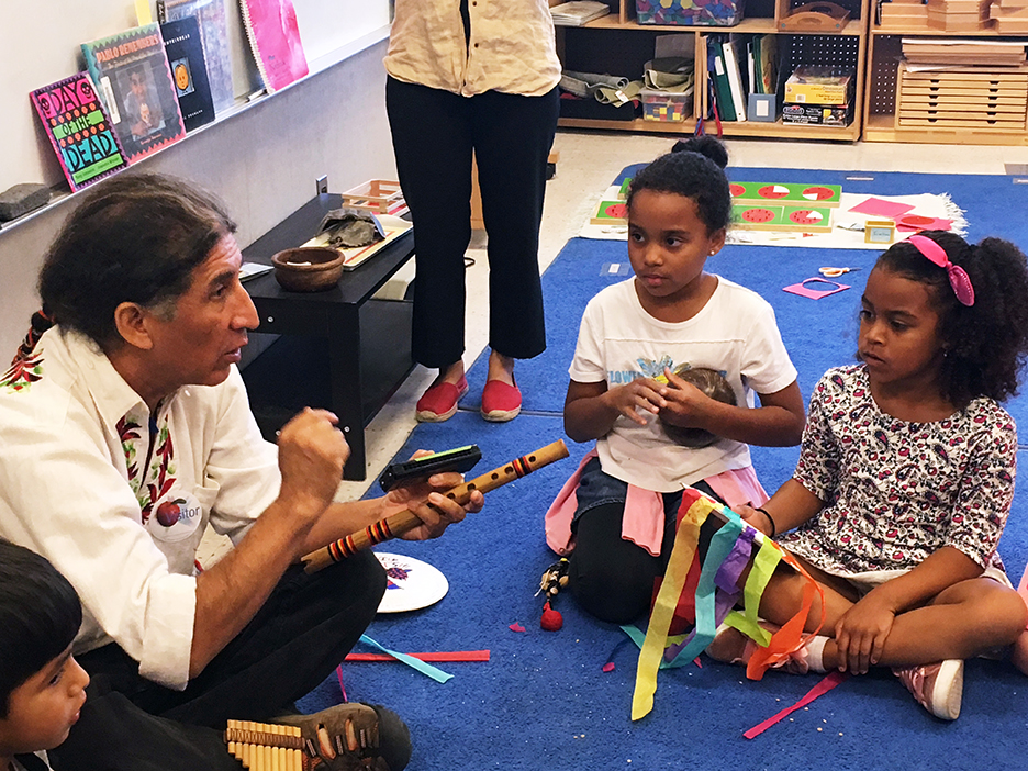 Native male shows instrument to two young female students of color while sitting on blue carpet