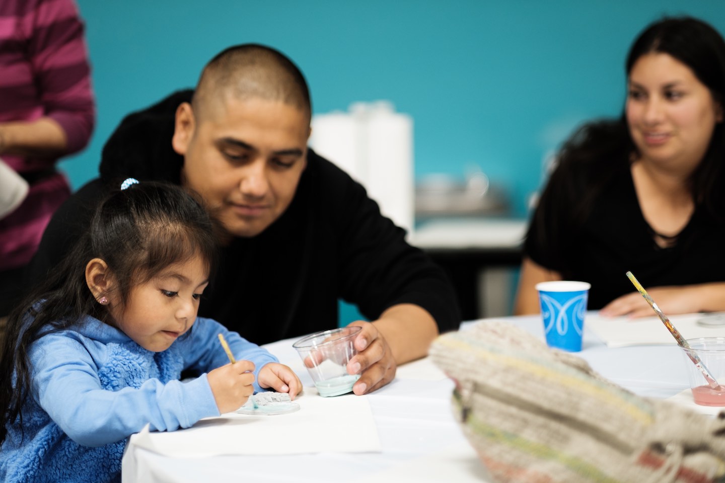 Latinx father and young daughter sit together while daughter paints, woman smiles and looks on in background