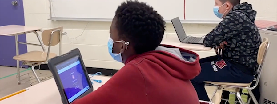Photo of student sitting at desk with laptop, wearing a mask