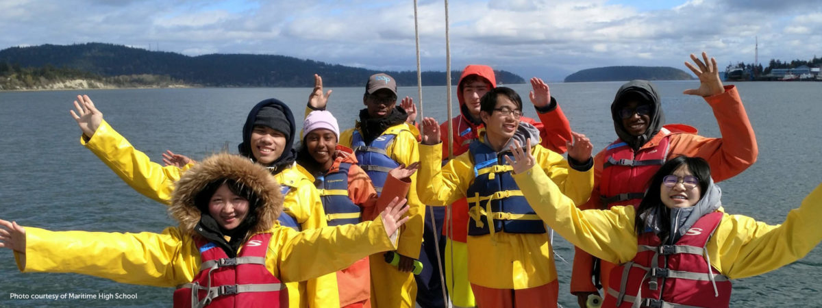 Diverse students wearing life jackets and yellow jackets smiling at camera in cold weather with water in the background