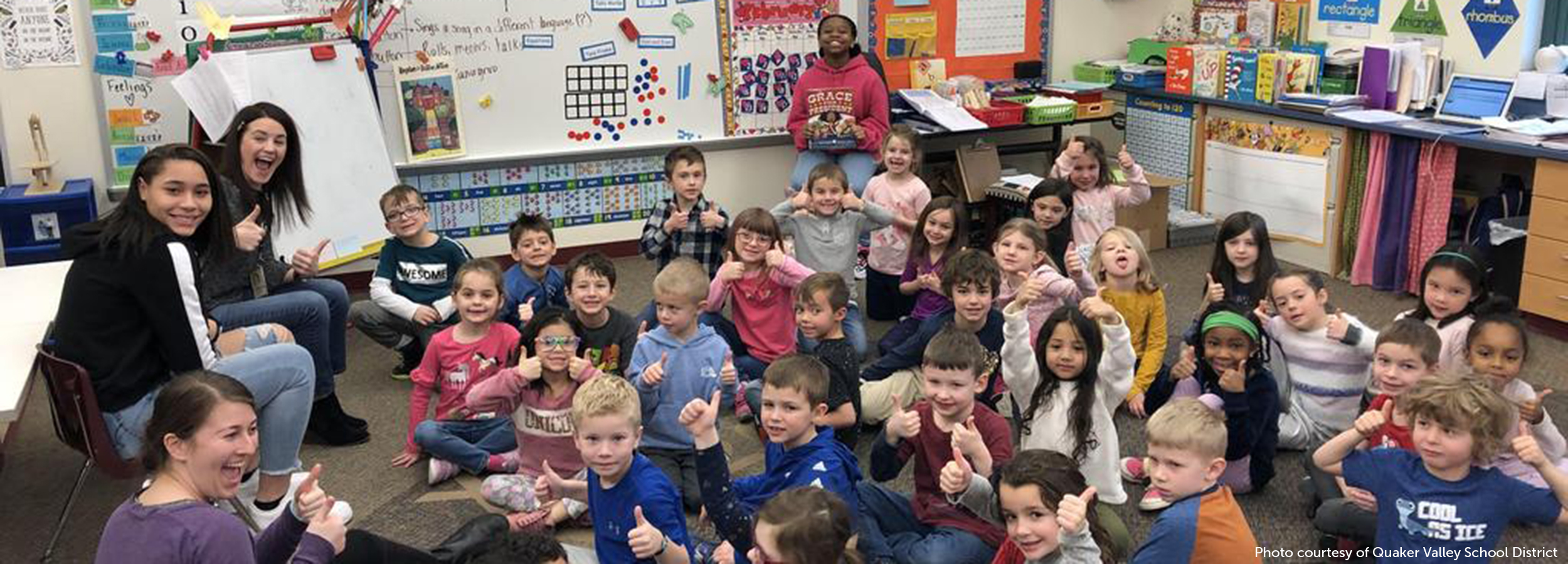 Photo from a Quaker Valley School District kindergarten classroom with students sitting on the floor all giving a thumbs up to the camera