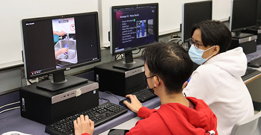 Two students sitting in front of computers, one looking at the other's screen which has a photo of hands filling a bottle in a sink.