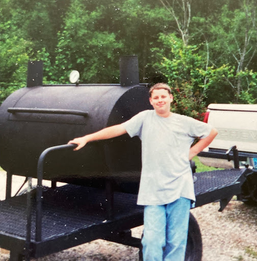 8th grade boy in T-shirt and jeans, smiling proudly in front of a meat smoker on wheels that is taller than him and hitched to a truck