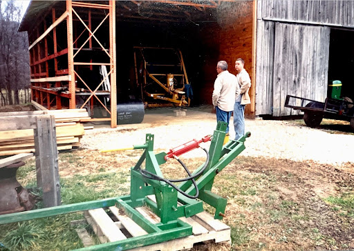 Father and son, with hands in pockets, looking at a barn shelf under construction. Foreground includes a green metal farm equipment.