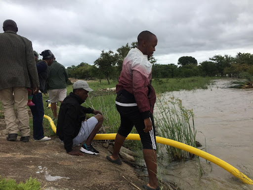 A group of people standing and bending down next to a large yellow irrigation tube dropped into a river.