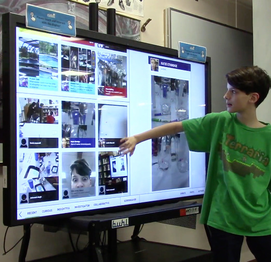 A student stands at a large computer screen. Displayed on the screen are images of science that have been contributed by the community. The student is scrolling through the images.