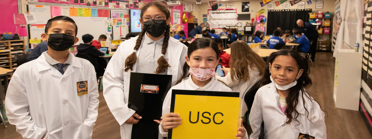 Four elementary students wearing white lab coats in a Compton Unified School District classroom smiling for the camera
