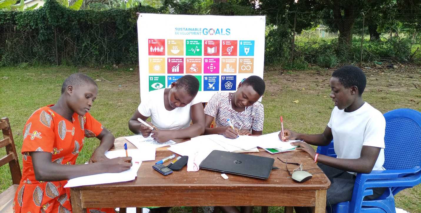 Four students sitting at an outside table and writing in notebooks, seated in front of a Sustainable Development Goals poster.