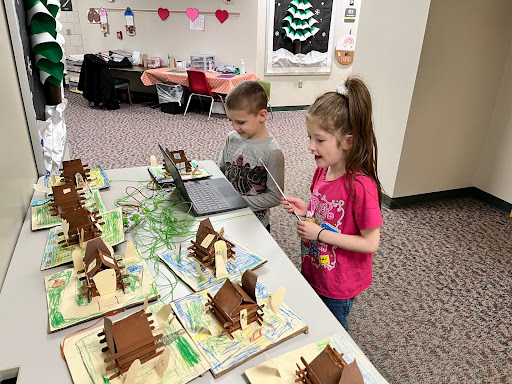 Two young students standing at a table looking at a laptop and a cardboard pioneer village that the students built and connected to the laptop for light and audio display. 