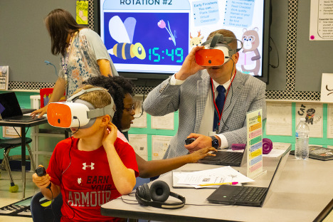 An adult man wearing an orange VR headset kneels next to a Black, female student sitting at a table as she controls a laptop. Another male student in the forefront also wears an orange VR headset and holds a controller.