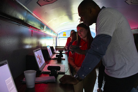A female student in a red shirt shows a Black adult male how to use software on a laptop inside of a revamped mobile virtual lab.
