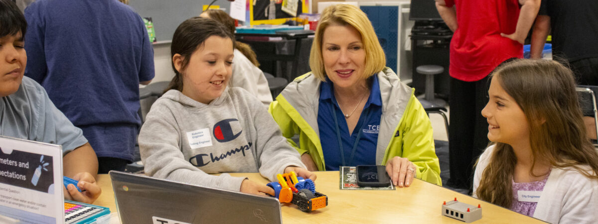 A teacher kneels next to a studeent who is showing off their robotics project