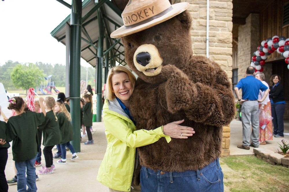 A white, adult woman hugs a brown bear mascot known as “Smokey Bear” outside of a school entrance. 
