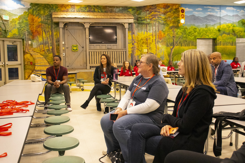 Five adults (three female and two male) sit in a cafeteria that has walls painted with forest and mountain landscapes, listening to a presentation from a speaker that is not visible in the photo. 