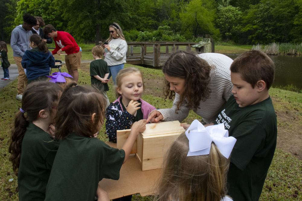 Four elementary-school aged children gather around a table outdoors to build a birdhouse. They are receiving directions from a young adult woman. 