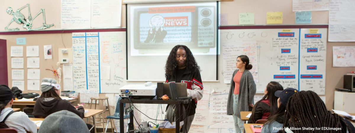 A high school student presents in front of her class. She is standing in front of a projection of an image of a television with a Breaking News banner