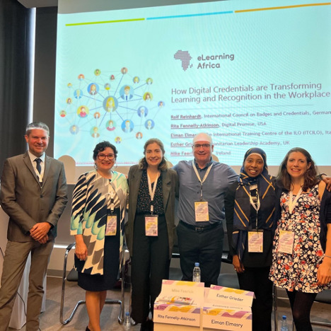 In order from left to right, Rolf Reinhardt, Rita Fennelly-Atkinson, April Williamson, Mike Feerick, Eiman Elmasry, and Esther Grieder pose for a photo in front of a slide reading “eLearning Africa: How Digital Credentials are Transforming Learning and Recognition in the Workplace.”