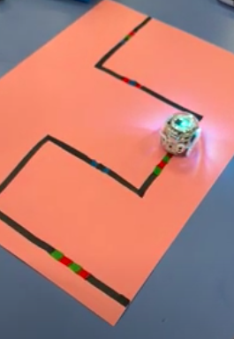 The image shows a drawing of a black line with a series of red, green and blue sections. The small circular robot is driving along the line with lights shining.