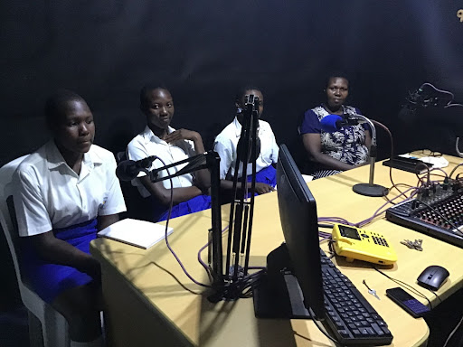 Educator and students sit at a table with microphones at a radio station. 