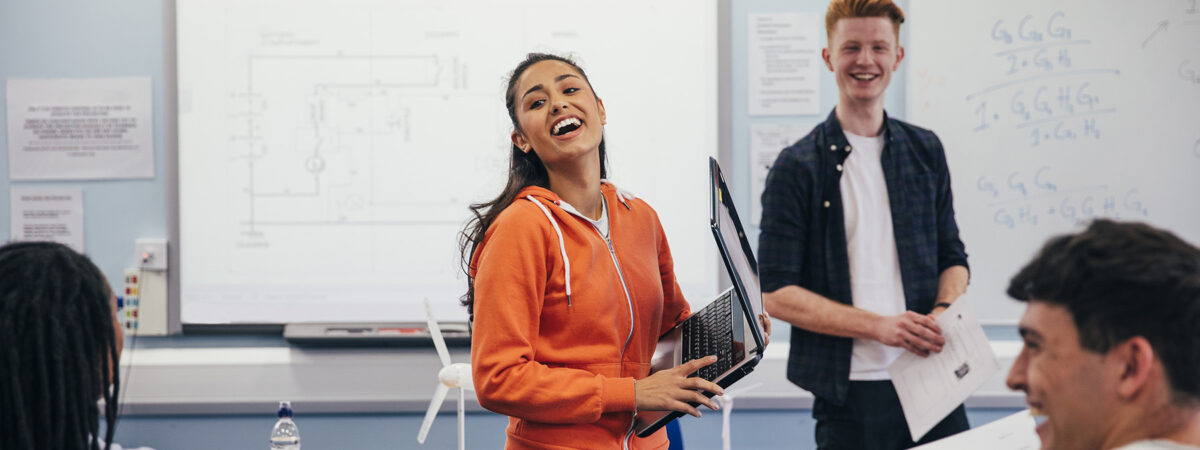 A young woman laughs and carries a laptop as she prepares to present her work to the class.