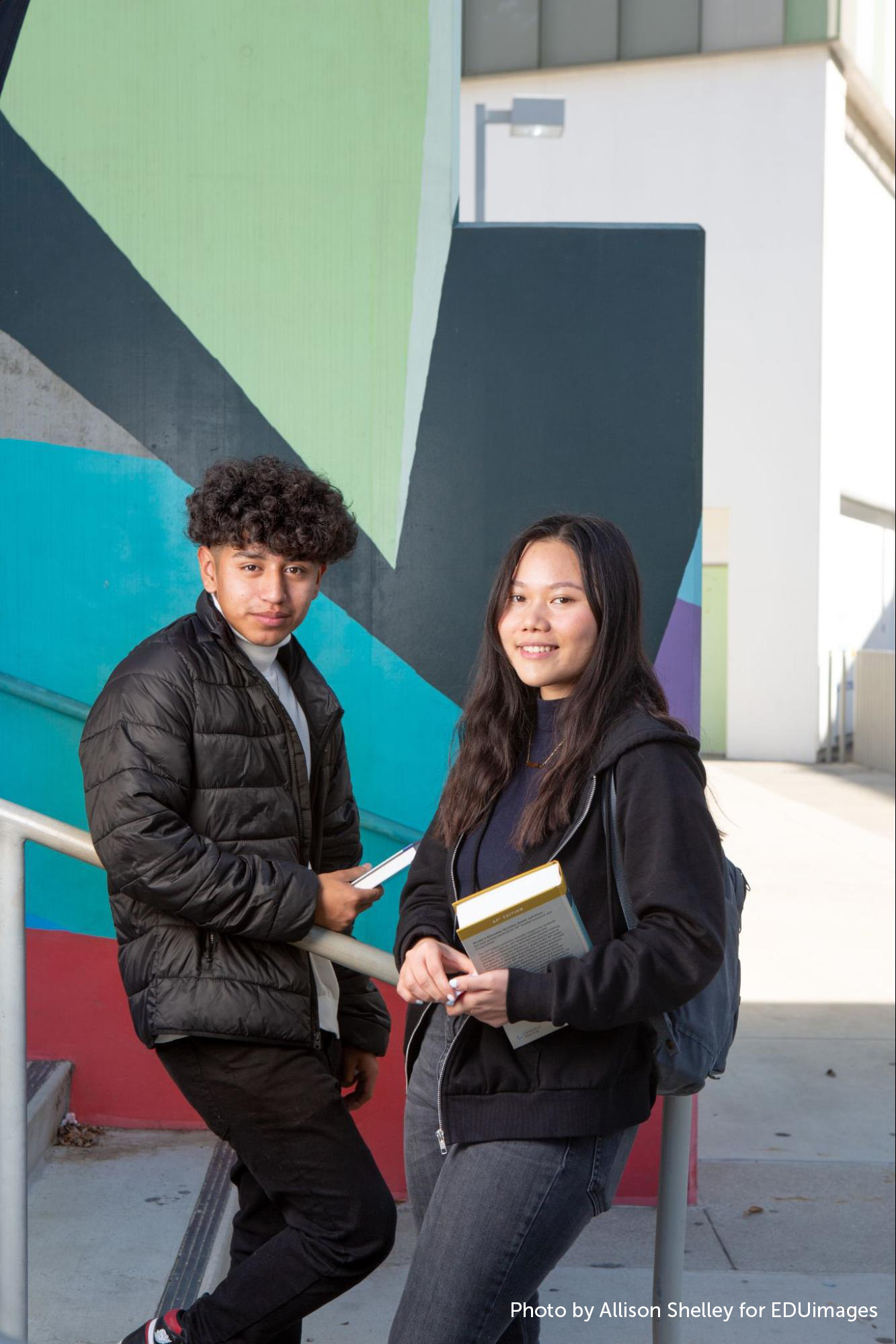 Two students, holding books, lean against the railing by the stairs outside their school