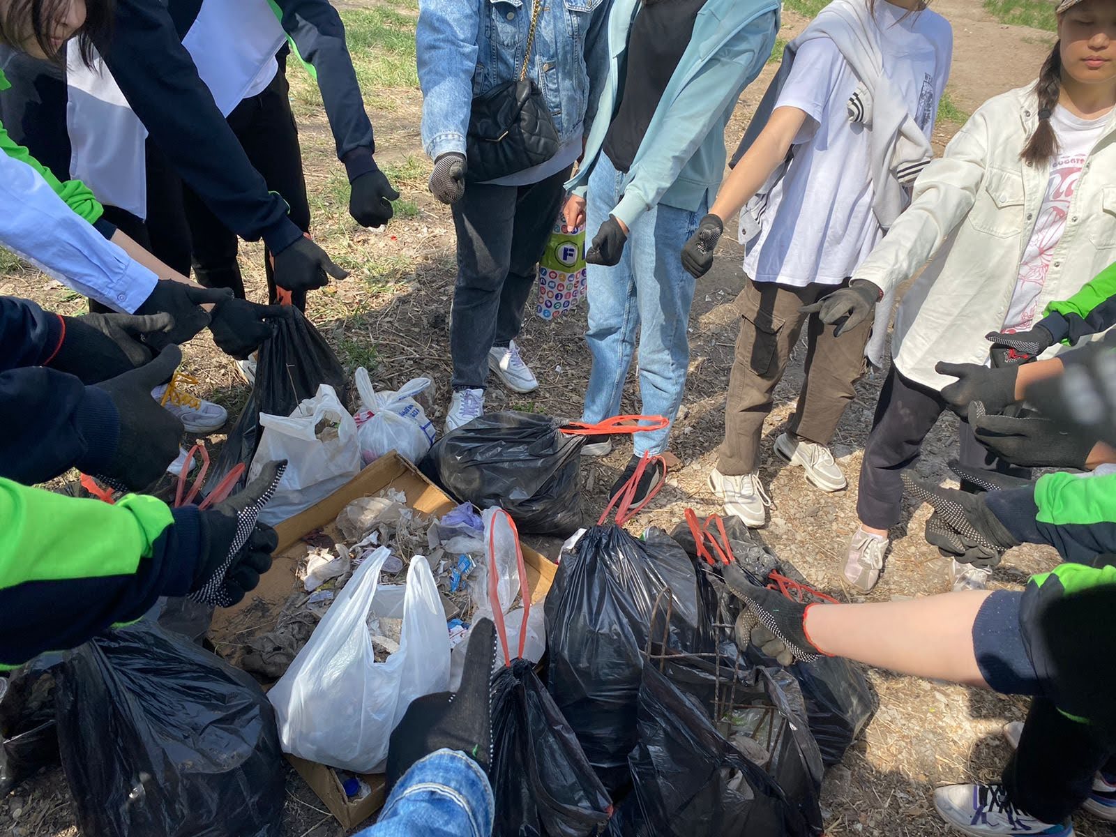 A pile of trash bags with recycled materials sits in the center of a circle of students who are standing around the pile with their fists reaching toward the center. 