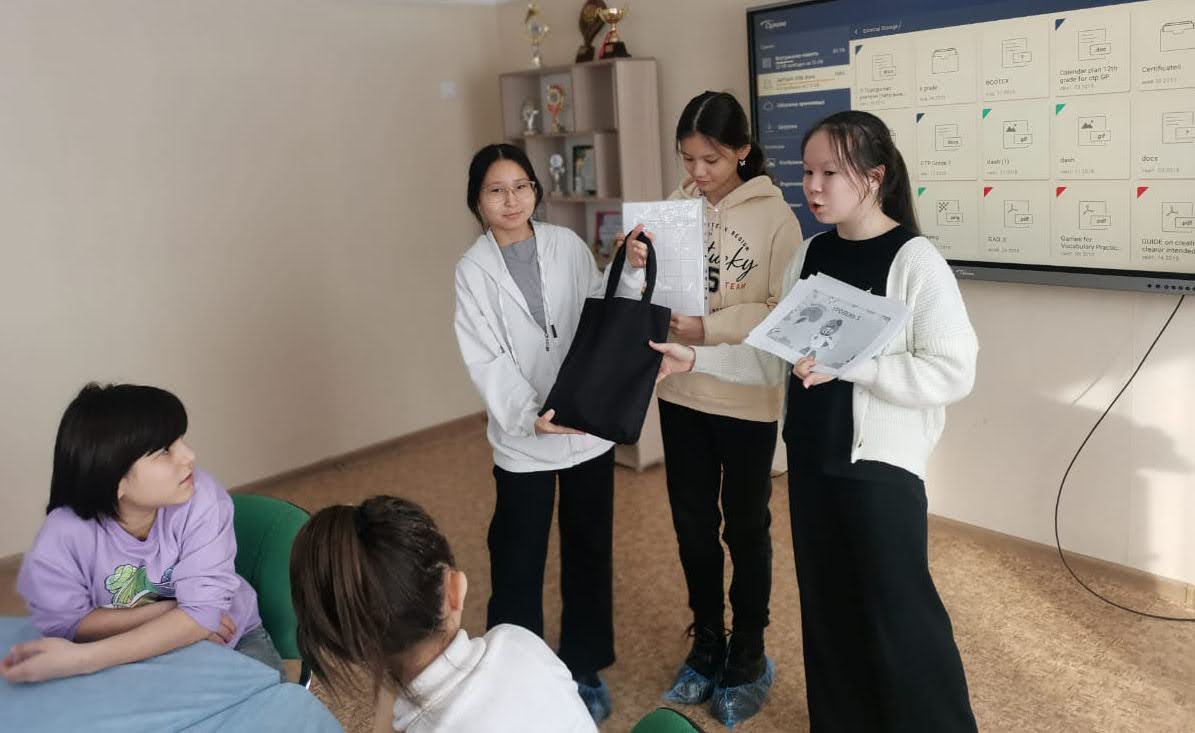 Three students present materials to a group of younger students in a classroom.