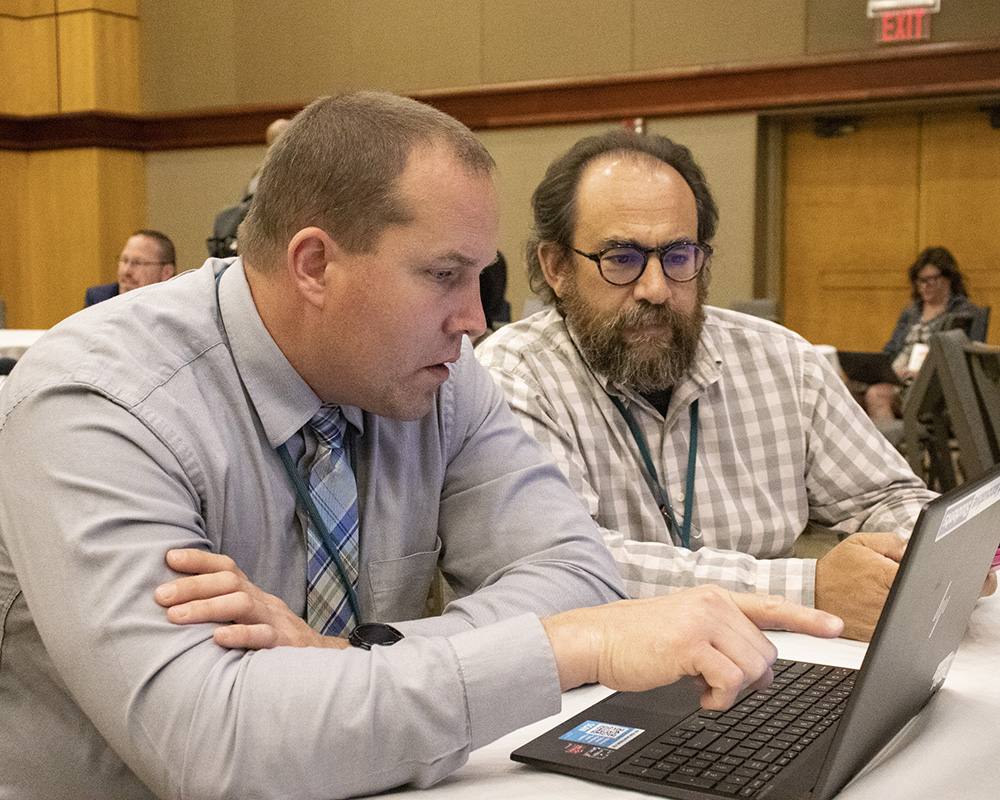 Two men sitting at a conference room table at a league convening looking at a laptop