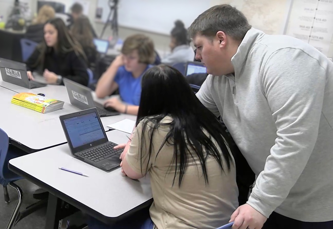 Coach Wallace stands to the right of a student and reaches to her computer. The computer has Net.Create with one of the table views showing. 