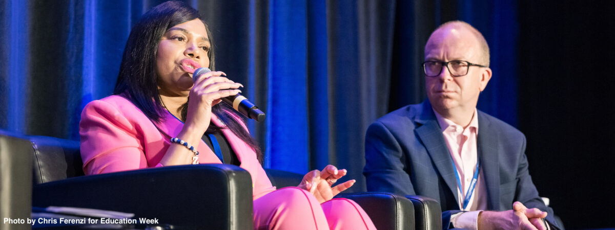 A black woman in a pink suit sits onstage and speaks into a microphone sitting next to a white man in a navy suit.